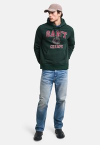 Sudadera verde oscura con el texto y logo "GANT CHAMPS", combinada con jeans de color azul claro y zapatos negros. El modelo está de pie con las manos en los bolsillos.