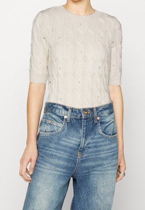 Kvinna klädd i en krämfärgad kabelstickad tröja med pärl- och rhinstenprydnader samt högmidjade bleka blå jeans.