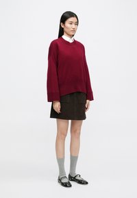 Burgundský pletený svetr s vykrojenými rameny, který je spárován s tmavohnědými sametovými sukněmi. Šedé ponožky a černé boty se stříbrnými akcenty dokončují vzhled.