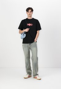 Svart t-skjorte med rød grafikk, lyseblå totebag, falmede denimjeans og hvite sneakers med røde striper. Modell som står foran en ensfarget bakgrunn.