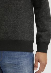 Schwarzes, strukturiertes Sweatshirt mit meliertem Finish, mit geripptem Saum und Bündchen. Getragen über blauen Jeans.