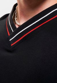 Black V-neck sweater with a smooth texture. Features red and white striped accents on the collar. Close-up view of the neckline and jewelry.