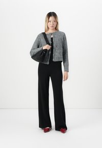 Woman wearing gray knit cardigan, black wide-leg pants, red pointed shoes, holding black shoulder bag against white background.