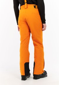 Persoon in feloranje skibroek met zwarte versterkte pijpen en zwarte skischoenen, staand tegen een witte achtergrond.