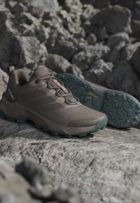 Brązowe buty trekkingowe Adidas Terrex z teksturowanymi zielonymi podeszwami umieszczone na skalnistym terenie na zewnątrz.