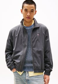 Giacca grigia con zip e colletto alto, polsini elasticizzati e logo. Indossata sopra un maglione blu con motivo e una camicia gialla. Jeans in denim chiaro.
