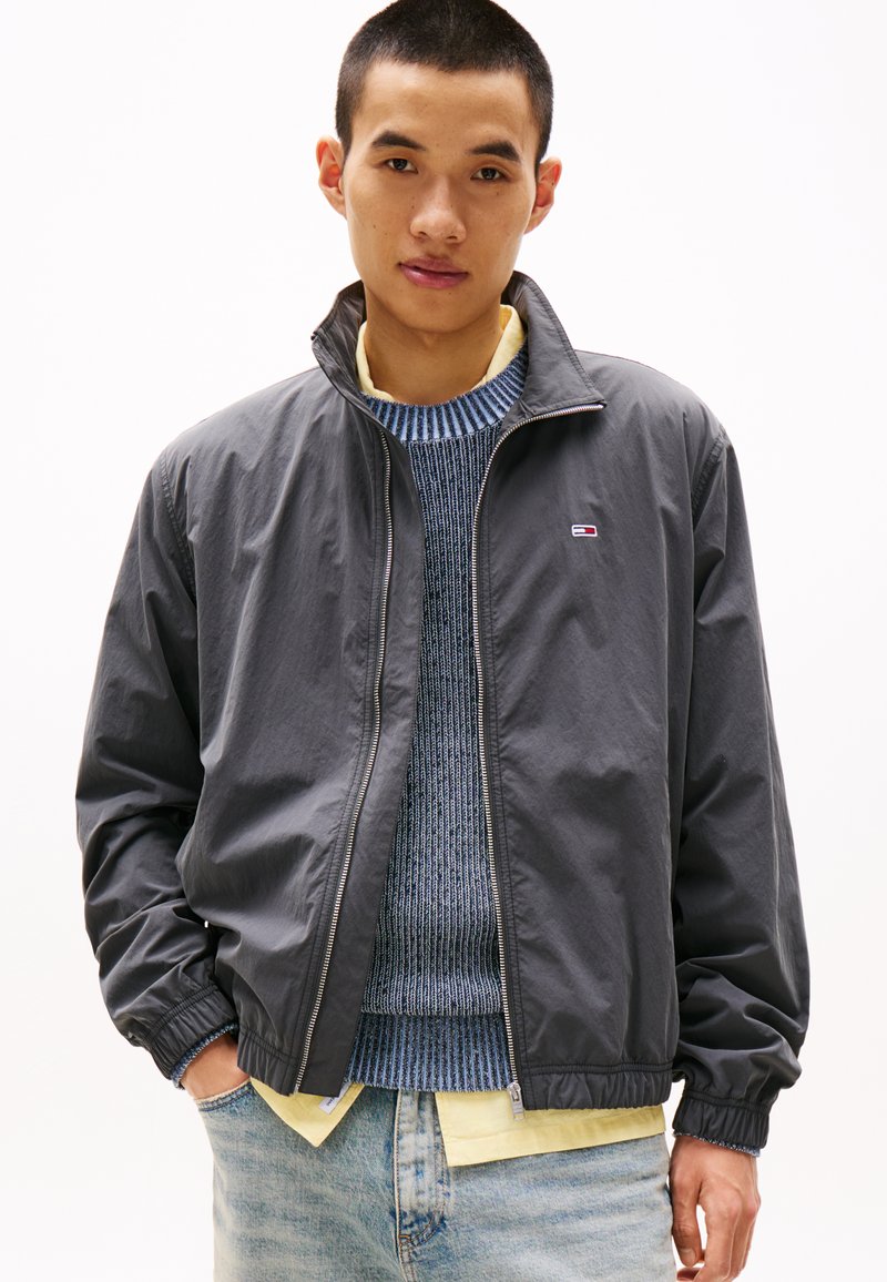 Giacca grigia con zip e colletto alto, polsini elasticizzati e logo. Indossata sopra un maglione blu con motivo e una camicia gialla. Jeans in denim chiaro.