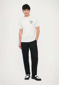 Bílé bavlněné tričko s grafickým motivem ve tvaru diamantu na levé straně hrudi, v kombinaci s černými kalhotami a černobílými botami.