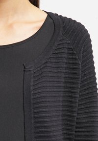 Schwarze Rippstrickjacke mit einem strukturierten Strickmuster, offener Vorderseite und gerundetem Ausschnitt, über einem einfarbigen schwarzen Oberteil getragen.