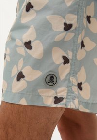 Shorts de bain bleu clair avec un motif floral en crème et marron foncé. Présente un bouton avec un design de tête de mort et tibias croisés.