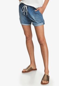 Shorts en denim avec une coupe décontractée, des revers roulés, une taille élastique avec cordon de serrage et des poches latérales. Associés à des sandales à glissière métalliques.