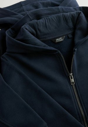 Námořnicky modrá mikina s kapucí z měkké látky, vybavená předním zipem, klokankovou kapsou a štítkem s názvem značky „NEXT“.