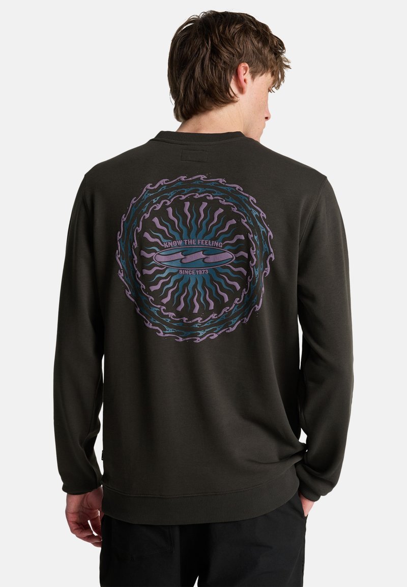 Hombre con sudadera negra que presenta un diseño de ondas circulares en púrpura y azul y un gráfico de una tabla de surf con la leyenda "Conoce la sensación desde 1973" en la espalda.