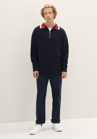Marineblauer Strickpullover mit Reißverschluss-Kragen, der mit einem rot-weißen Streifenakzent versehen ist. Kombiniert mit marineblauen Hosen und weißen Sneakers.