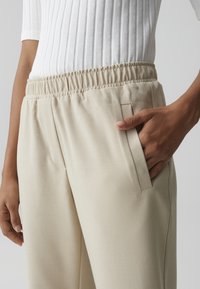 Persona che indossa pantaloni beige con vita elastica e una mano nella tasca anteriore, abbinati a una maglia bianca a coste.