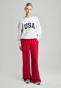 Harmaa huppari, jossa on "USA" graafinen kuva tummansinisellä ja vaaleansinisellä, yhdistettynä punaisiin leveälahkeisiin housuihin. Mallilla on päällään vaaleat lenkkarit. Yksinkertainen tausta.