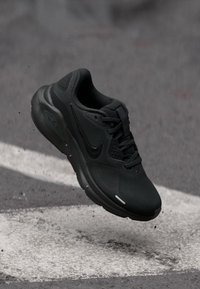 Chaussure de course Nike noire en l'air au-dessus de l'asphalte avec des marquages routiers blancs, suggérant du mouvement ou un saut.