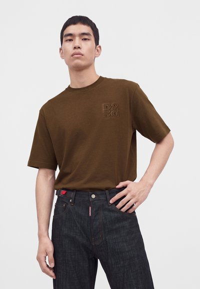 Jeune homme portant une chemise marron à manches courtes avec un logo brodé et un jean en denim foncé, debout avec une main sur la hanche.