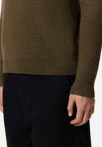 Olivgrön ribbad stickad tröja med åtsittande fåll och långa ärmar, kombinerad med svarta byxor. Texturerade tygdetaljer syns.