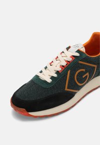 Sneaker verde e nera con texture a zig zag, accenti arancioni e lacci crema. Logo notevole in arancione sul lato.