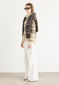 Pull en maille épaisse beige avec des motifs géométriques multicolores, porté avec un pantalon blanc à jambes larges et des chaussures à talons beiges pointues. Lunettes de soleil noires.