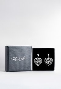 Silverfärgade hjärtformade örhängen med intrikata utskärningar, presenterade i en svart sammetfodrad låda med en texturerad svart yta.