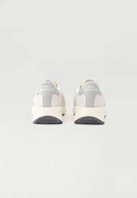 Vita och ljusgrå sneakers med en texturerad ovandel, vadderad krage och gummisula, med "New Balance"-logotyp på hälen och sidan.