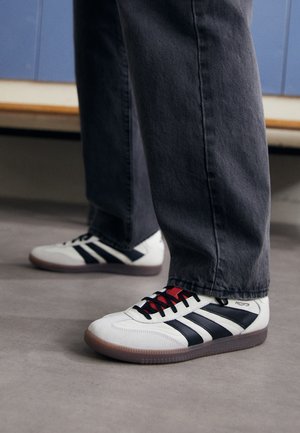 Hvide sneakers med sorte striber og røde snørebånd, båret med mørke bukser på gråt gulv, indendørs nær blå skabe.