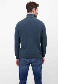 Granatowy dzianinowy sweter z wysokim kołnierzem, ściągaczami na mankietach i na dole oraz teksturowanym wzorem. Noszony z jeansami o średnim odcieniu.