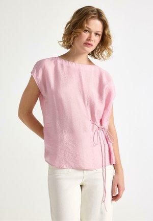 Kvinde iført en løstsiddende, skinnende lyserød bluse med sidetie-detalje og hvide bukser, stående mod en ensfarvet lys baggrund.