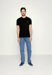 T-shirt preta de mangas curtas com um pequeno logótipo vermelho, combinada com jeans azul claro e ténis azul-marinho. Modelo posicionado centralmente, em pé e ereto.