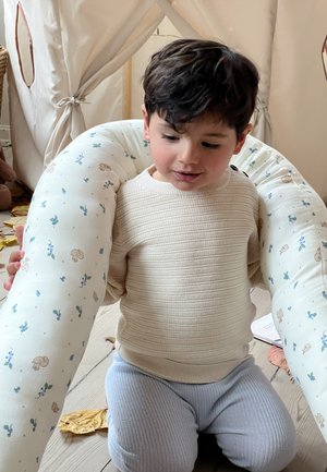 Hvid, plys amme pude med blå blomster- og dyremønstre; et barn i en struktureret beige sweater sidder tæt på, bløde grå bukser.