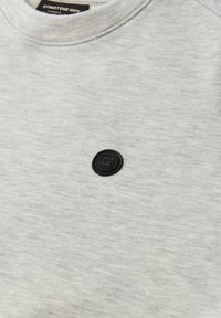 Lysegrå crewneck-trøje med et lille rundt sort emblem med et geometrisk design nær kraveområdet ved brystet.