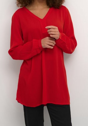 Femme portant un chemisier rouge à manches longues avec col en V, tissu texturé et coupe ample, associée à un pantalon noir, debout devant un fond blanc.