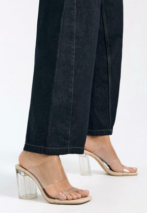 Feet wearing clear, open-toe block heels paired with dark wide-leg denim pants against a plain white background.