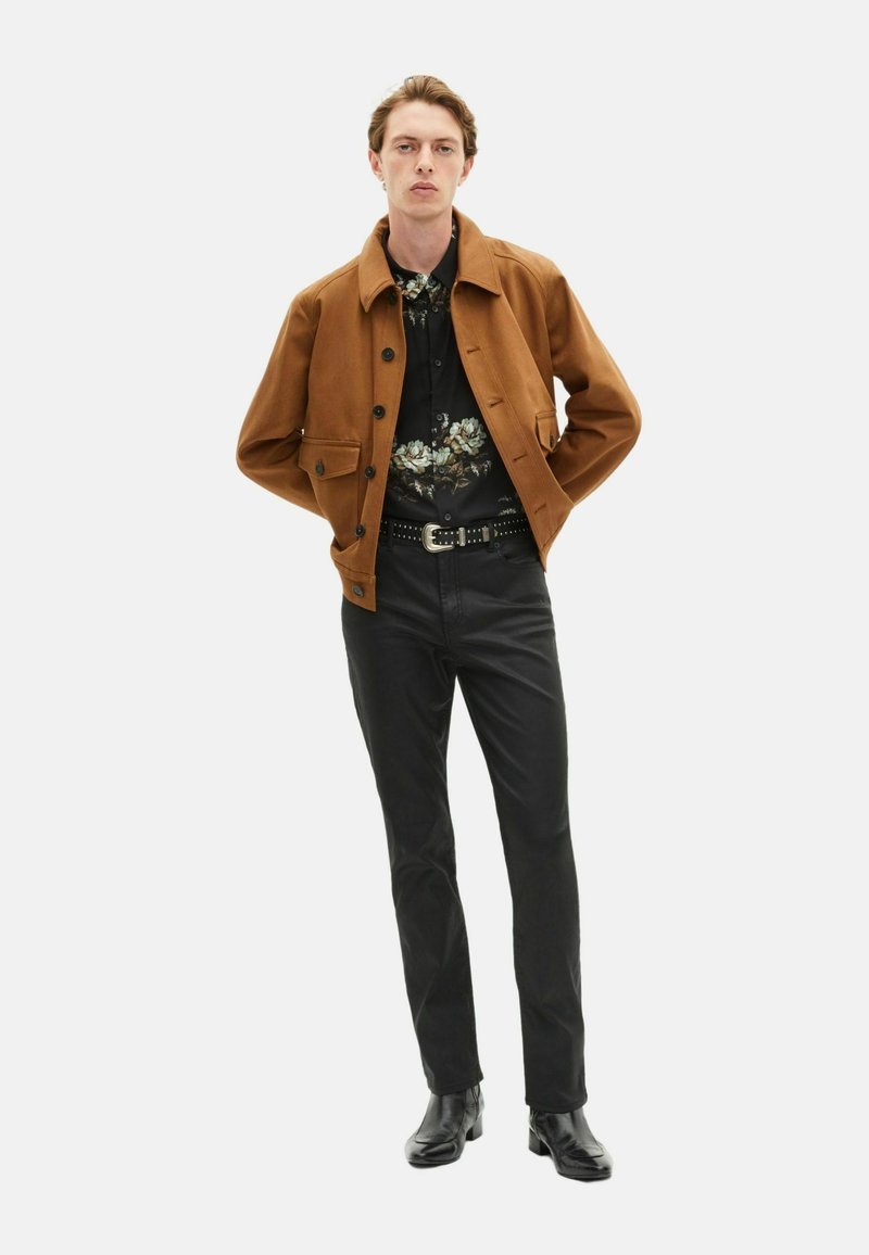 Veste marron avec un col et des boutons, portée sur une chemise noire à fleurs, assortie avec un pantalon noir et des bottes noires. Textures lisses dans l'ensemble.