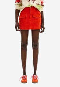 Jupe en denim rouge avec une taille ajustée, deux poches avant et une fermeture à bouton. Associée à des baskets orange vif avec des lacets roses.