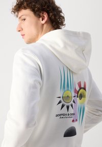 Valge kapuutsiga särk, millel on seljal graafiline disain abstraktsete silmade ja päikesepaiste mustritega kollases, teal'ides, punases ja mustas.