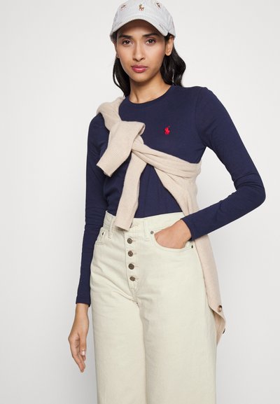 Granatowa bluzka z długim rękawem z czerwonym logo, beżowy sweter z dzianiny przewiązany w talii oraz kremowe spodnie z wysokim stanem z detalem w postaci guzików.