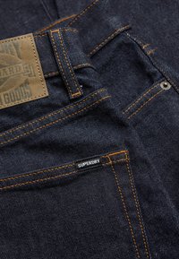 Primer plano de unos vaqueros de mezclilla azul oscuro con costuras naranjas, un bolsillo delantero con una etiqueta "Superdry" y un parche marrón de la marca en la cinturilla.