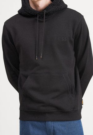 Czarna bluza z kapturem wyściełanym teksturowanym materiałem sherpa, sznurkami oraz przednią kieszenią typu kangur, noszona przez osobę w niebieskich spodniach.