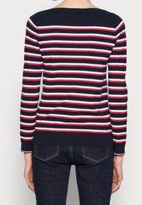 Marineblaues Langarmshirt mit horizontalen roten und weißen Streifen, Rundhalsausschnitt und gerippten Bündchen und Saum; getragen mit dunklen Jeans.