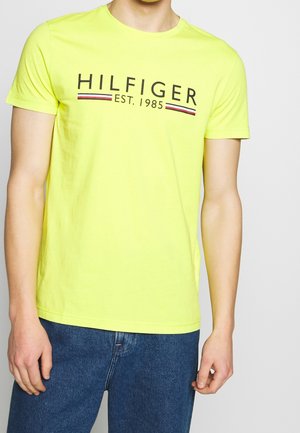 Personne portant un T-shirt jaune à manches courtes avec le texte "HILFIGER EST. 1985" et un jean bleu, sur un fond clair unicolore.