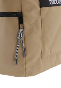 Mochila de nailon beige con cremallera negra, que presenta un cordón de tiro a rayas y un diseño elegante, mostrando costuras geométricas y una superficie texturizada.