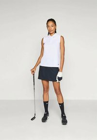 Ženska stoji s golf palico, oblečena v belo brezrokavno polo majico, črno krilo, črne nogavice, golf rokavico in črne superge na enobarvni podlagi.