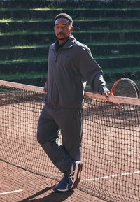Mann i grå treningsdrakt som hviler seg på tennisnettet, holder en rød og svart tennisracket, står på leirbane med grønne trinn i bakgrunnen.