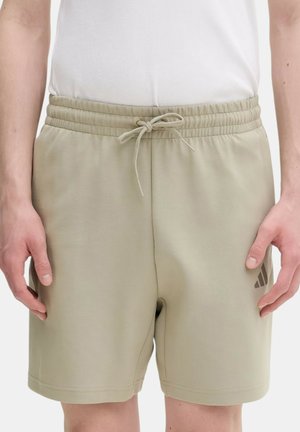 Uomo che indossa pantaloncini beige con vita elasticizzata e coulisse con un piccolo logo sulla gamba sinistra, mani appoggiate ai fianchi, camicia bianca infilata.