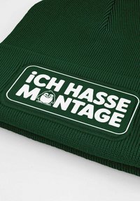 Dark green beanie featuring a ribbed texture and a white rectangular patch with the text "ICH HASSE MONTAGE" and a cartoon character.