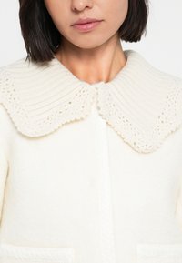 Κρεμ μάλλινη ζακέτα με υφασμένο, πλεκτό γιακά. Διαθέτει κούμπωμα με κουμπιά και δύο μπροστινές τσέπες. Μαλακό, ζεστό ύφασμα.