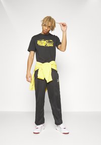 Sort grafisk T-shirt med gul print, parret med sorte bukser og en bundet gul sweatshirt. Fodtøj: hvide sneakers med røde accenter.